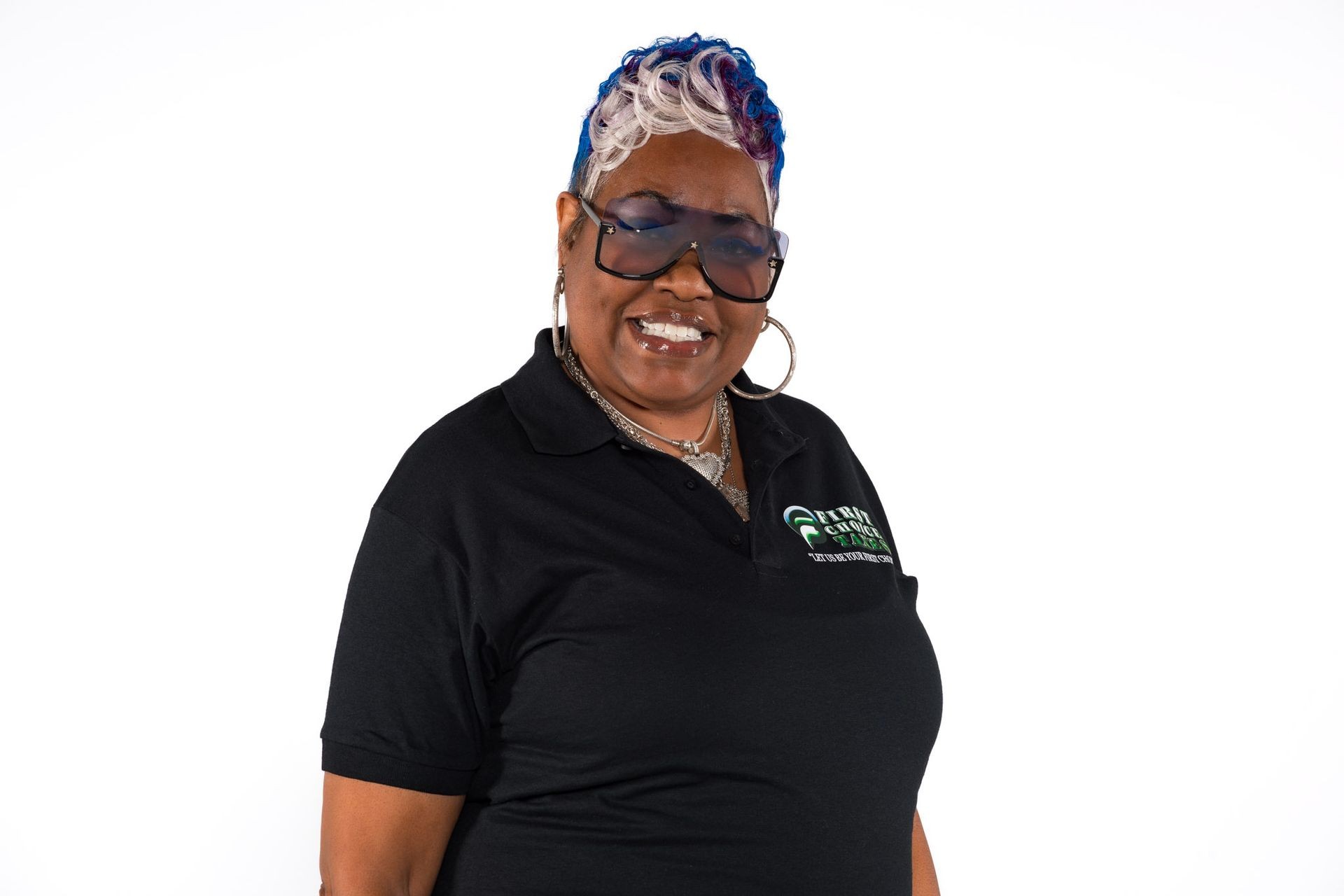 Woman in black polo shirt with bold sunglasses and colorful hair, smiling against a white background.